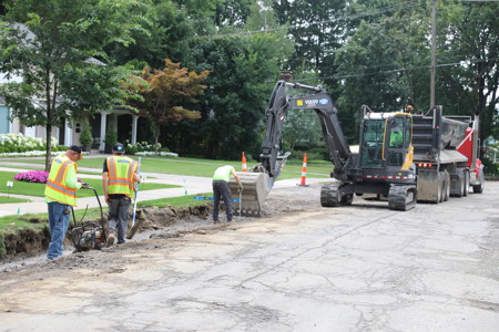 Road crew workers use equipment and an excavator to work on crumbling neighborhood roads
