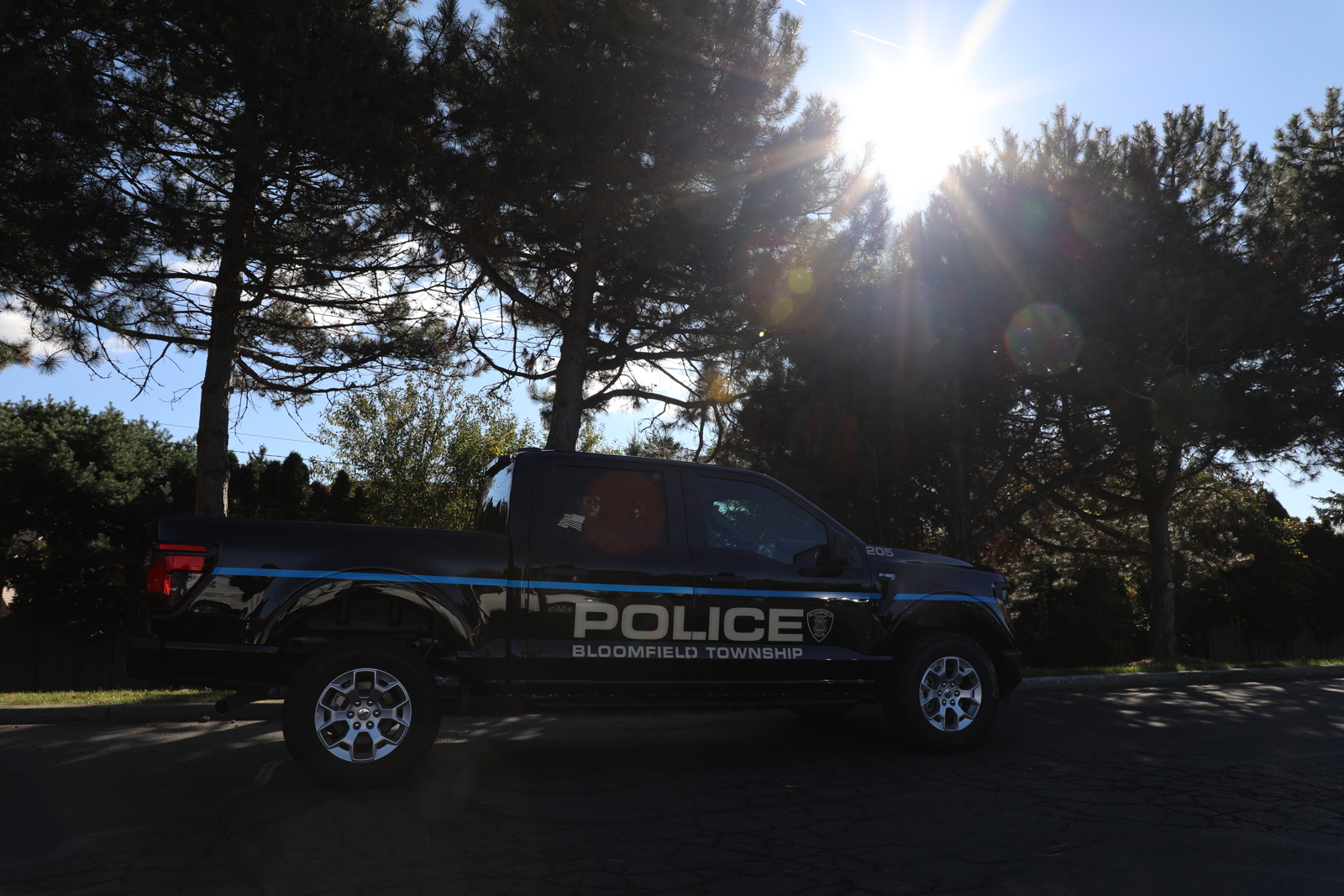 Dark shiny pick up truck that reads Police Bloomfield Township on the side parked next to tall pine trees in the shadows beneath a sunny blue sky.