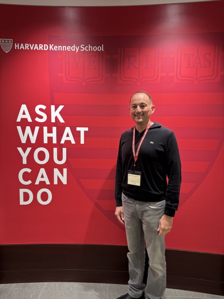 Michael Shostak standing in front of a wall with the quote "Ask What You Can Do" and the logo for Harvard Kennedy School