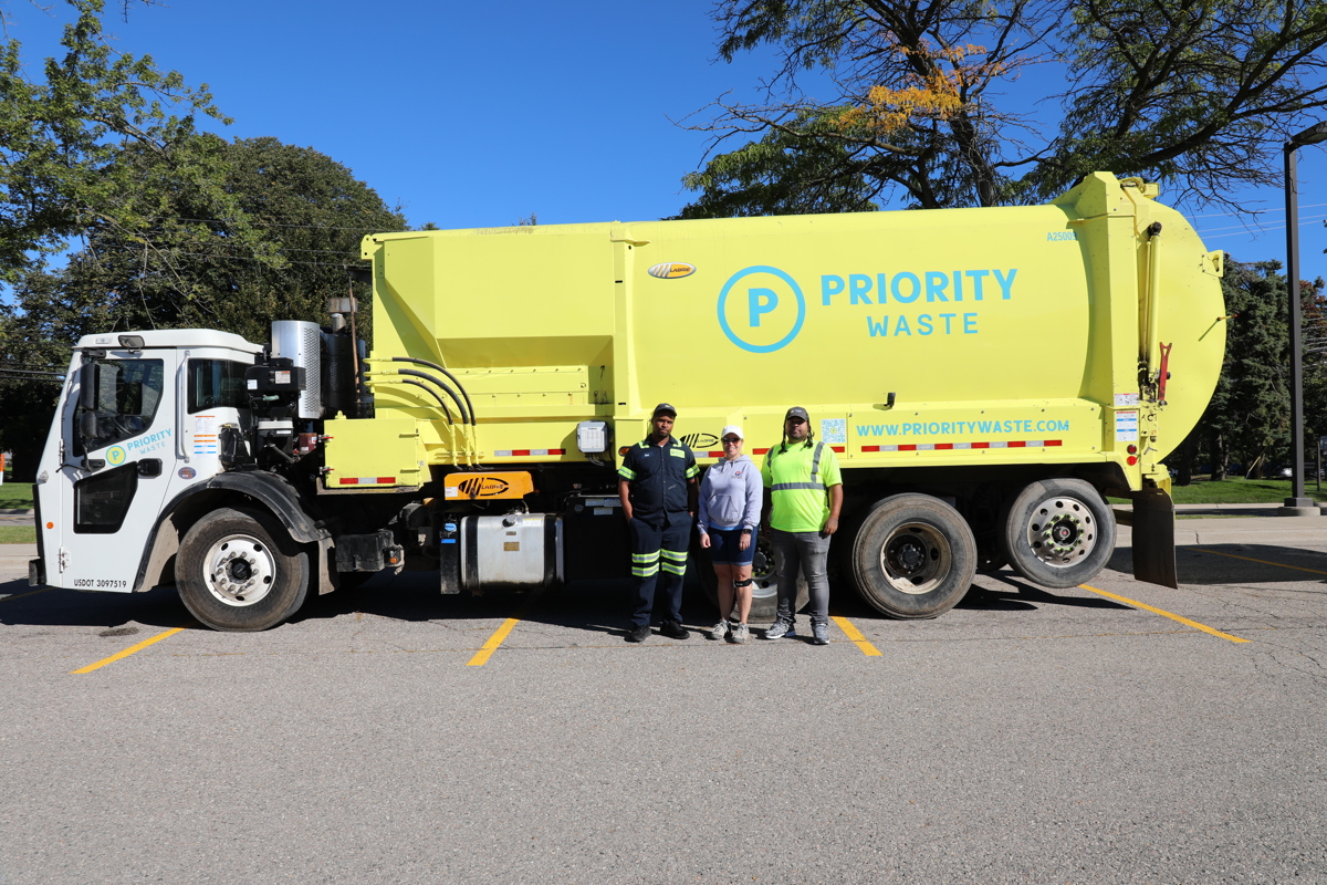 A new yellow Priority Waste garbage truck with 3 workers standing along side it.