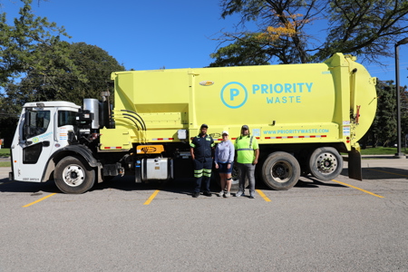 A new yellow Priority Waste garbage truck with 3 workers standing along side it.
