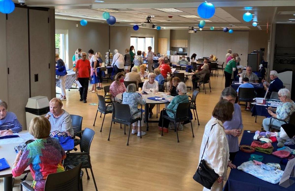 large gathering room full of people sitting at round tables and perusing rectangular tables along the perimeter. Room looks festive with blue balloons hanging from ceiling.