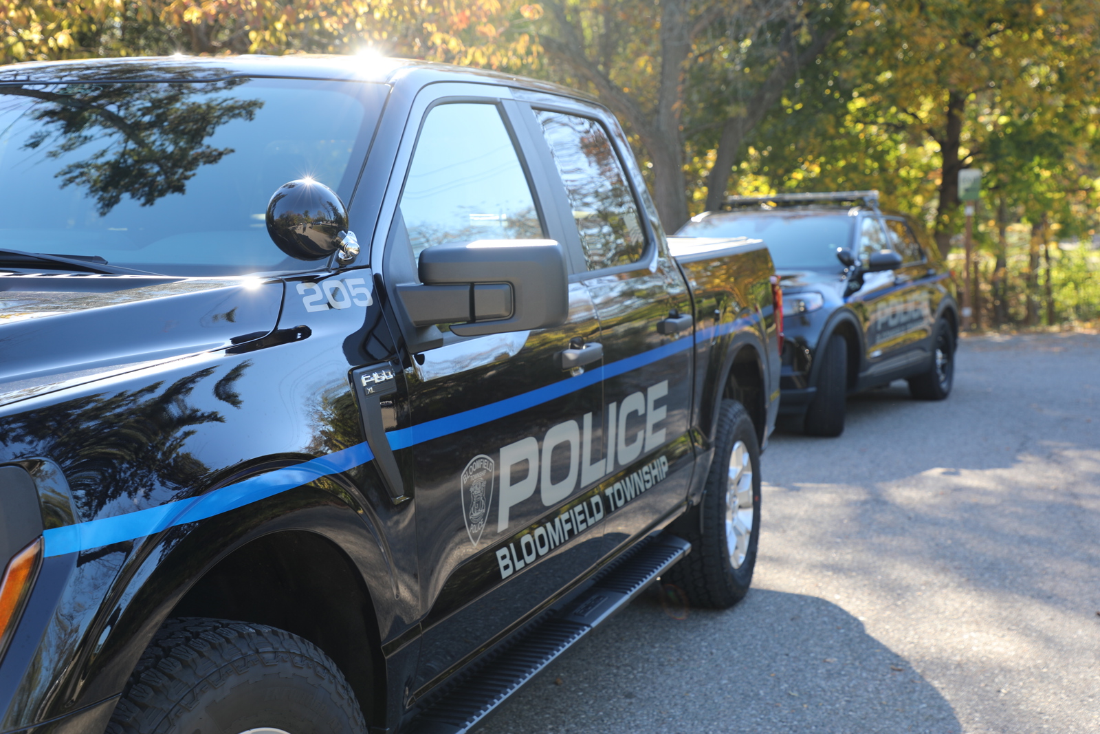A dark Bloomfield Township Police pickup truck parked on pavement next to green trees with a patrol card of same color and same logo-wording parked behind it.
